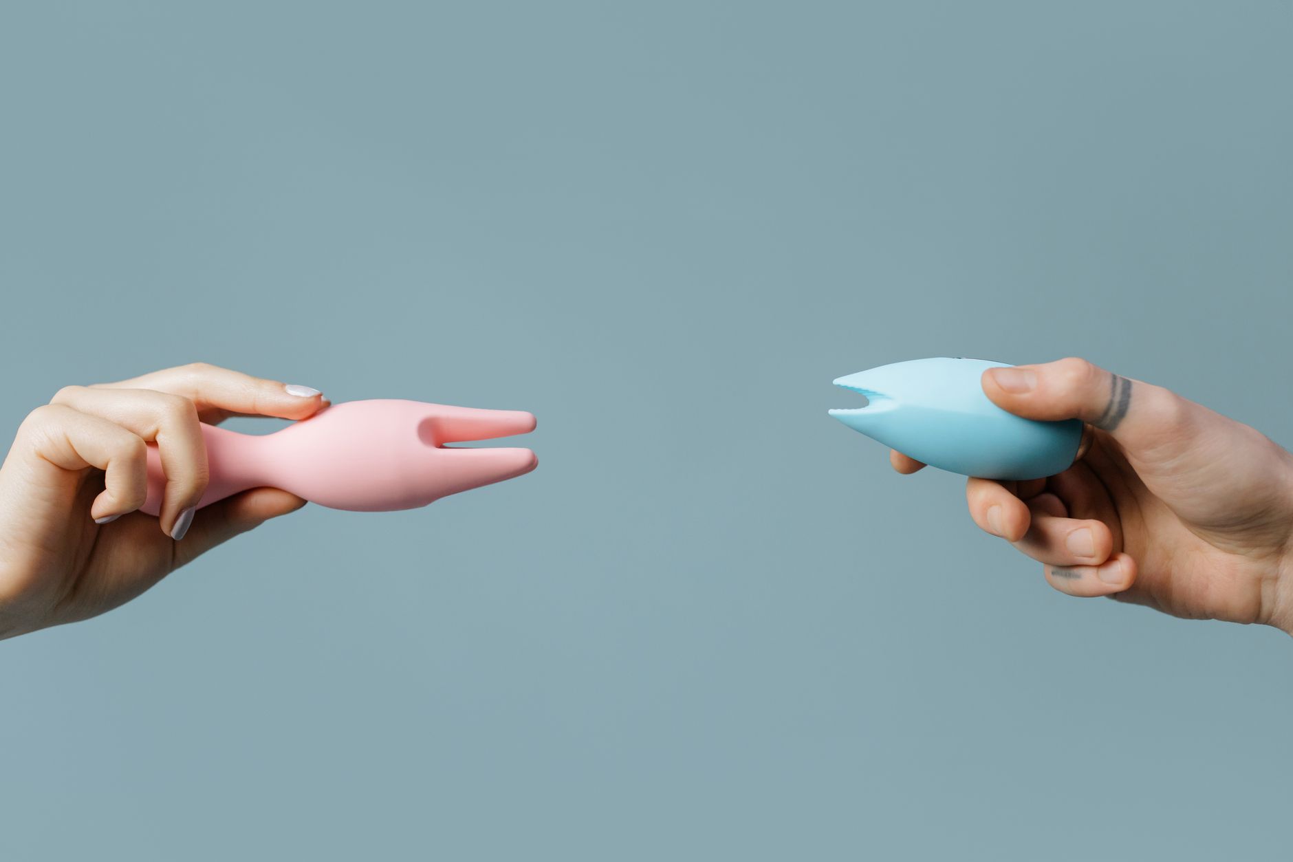 Two hands holding silicone vibrators in complementary colors against a soft pastel background